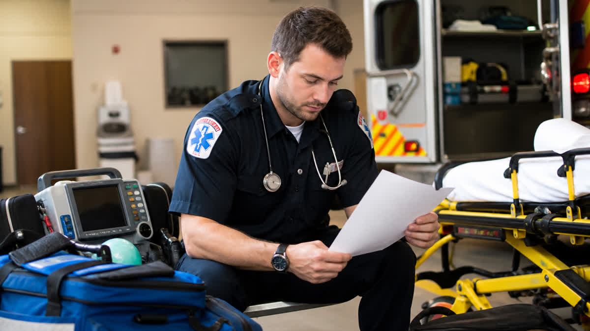 EMT reviewing a resume beside ambulance stretcher blood pressure cuff and portable cardiac monitor in a fire-EMS station bay