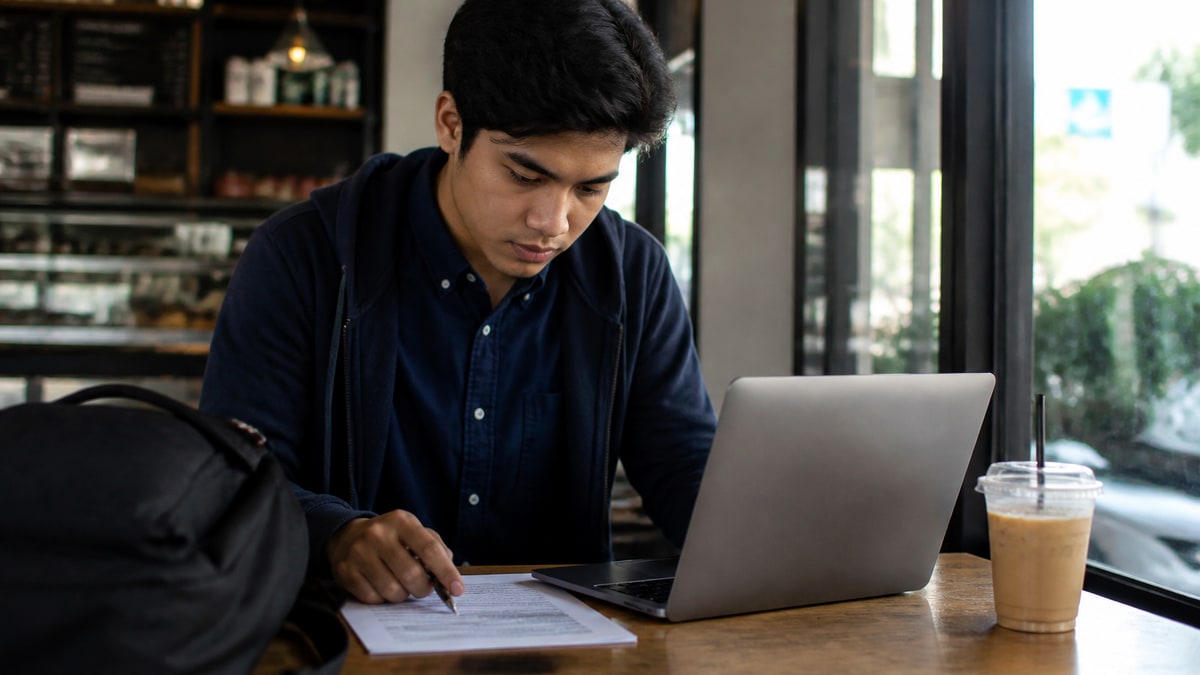 Recent graduate reviewing a one-page resume and laptop job application at a coffee shop table